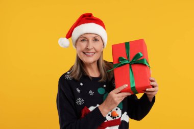 Noel süveteri ve turuncu arka planda Noel Baba şapkası tutan mutlu son sınıf kadını.