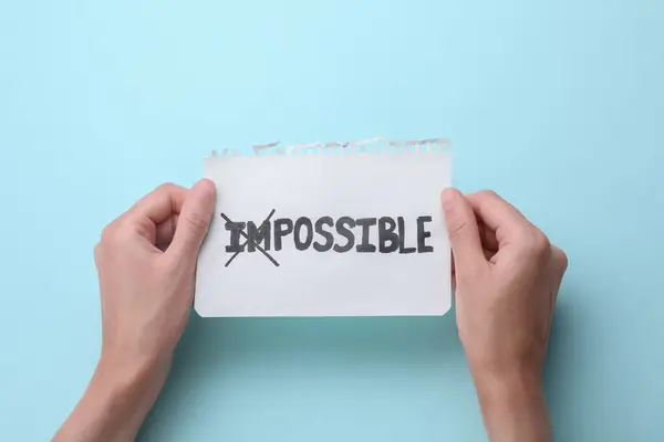 Motivation concept. Woman holding paper with changed word from Impossible into Possible by crossing over letters I and M on light blue background, top view