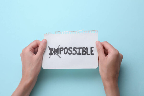 Motivation concept. Woman holding paper with changed word from Impossible into Possible by crossing over letters I and M on light blue background, top view