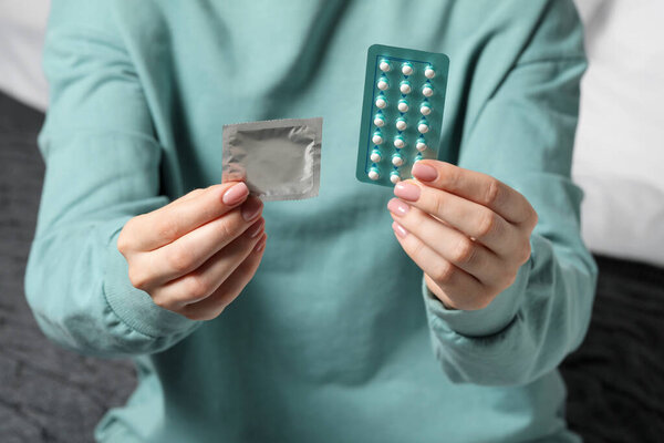 Woman holding condom and contraceptive pills on blurred background, closeup. Choosing birth control method