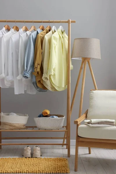Wardrobe organization. Rack with different stylish clothes, armchair and lamp near grey wall indoors