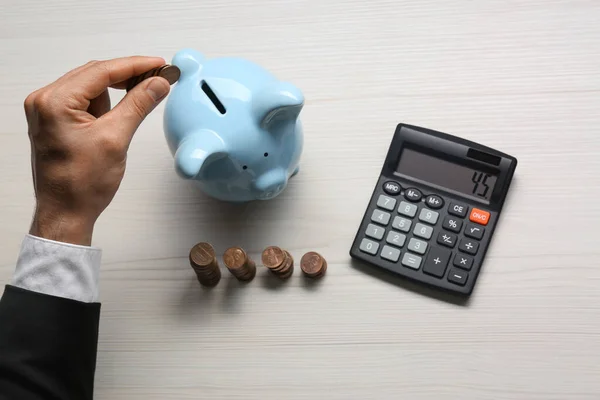 Budget planning. Businessman putting coin into piggy bank at light wooden table, top view