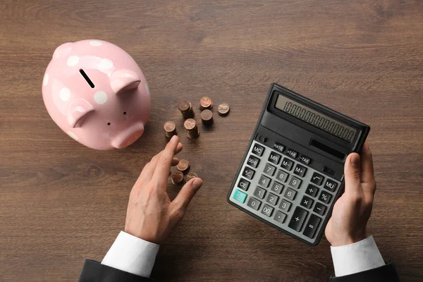 Budget planning. Businessman with piggy bank calculating at wooden table, top view
