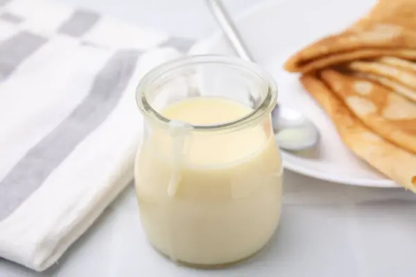 Tasty condensed milk in jar and crepes on white table, closeup