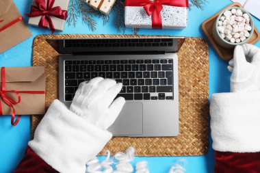 Noel Baba dizüstü bilgisayar kullanıyor, yakın plan. Hediye kutuları, bir fincan sıcak içecek ve açık mavi arka planda Noel dekoru.