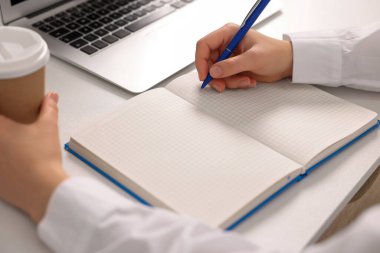 Beyaz masadaki deftere bir fincan kahve yazan genç kadın, yakın plan.