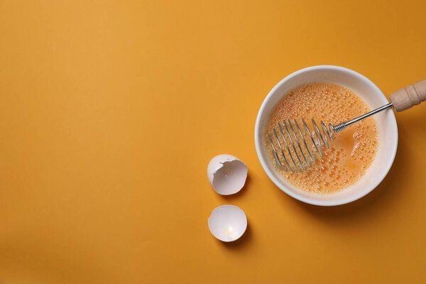 Beaten eggs, metal whisk in bowl and shells on orange background, flat lay. Space for text
