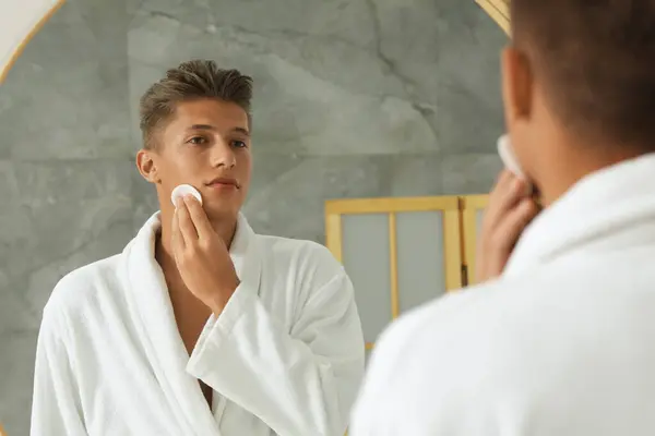 浴室の鏡の近くにコットンパッドで顔を掃除するハンサムな若者 — ストック写真