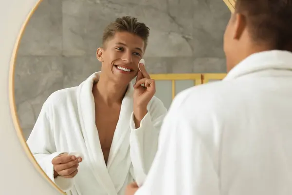 浴室の鏡の近くにコットンパッドで顔を掃除するハンサムな若者 — ストック写真