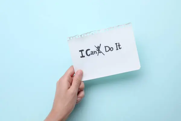 Motivation concept. Woman holding paper with changed phrase from I Can\'t Do It into I Can Do It by crossing over letter T on light blue background, top view