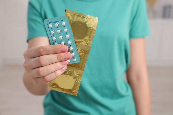 Woman holding condoms and contraceptive pills on blurred background, closeup. Choosing birth control method