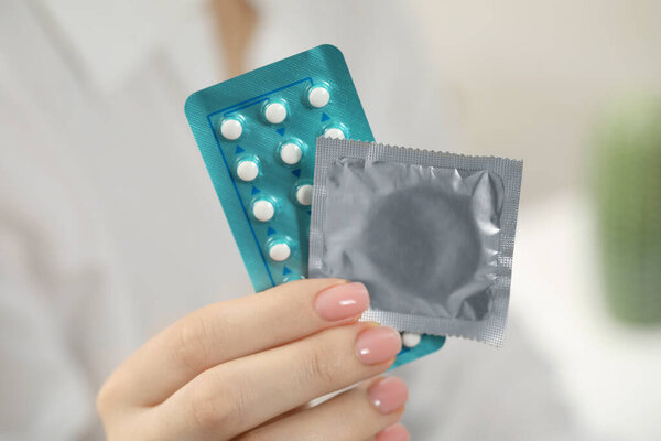 Woman holding condom and contraceptive pills on blurred background, closeup. Choosing birth control method