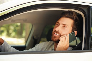 Mutlu sakallı adam arabada akıllı telefondan konuşuyor, dışarıdan bakıyor.
