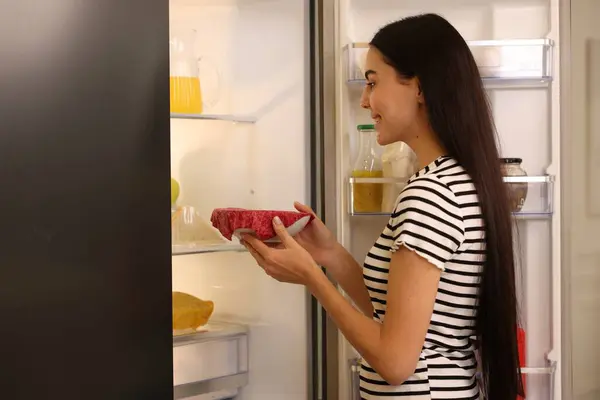 Woman putting bowl covered with beeswax food wrap into refrigerator indoors