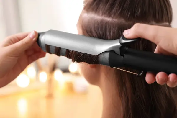 Hairdresser curling woman's hair with iron in salon, closeup
