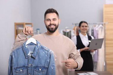 Kuru temizleme servisi. İçeride plastik torbada kot ceketli, askılık tutan mutlu bir adam. İşyerinde dizüstü bilgisayar kullanan işçiler