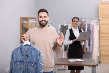Kuru temizleme servisi. Kot ceketli, askılık tutan mutlu bir adam ve içeride iyi bir jest gösteriyor. İşyerinde dizüstü bilgisayar kullanan işçiler
