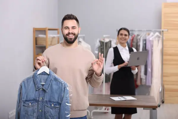 Dry-cleaning service. Happy man holding hanger with denim jacket and showing ok gesture indoors. Worker using laptop at workplace