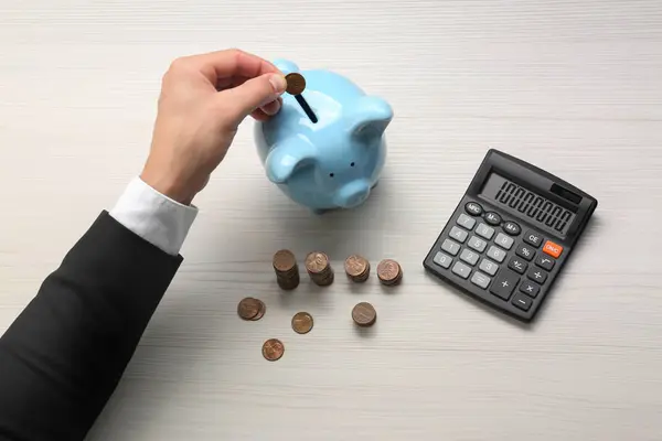 Budget planning. Businessman putting coin into piggy bank at light wooden table, closeup