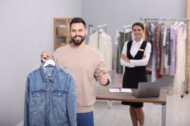Kuru temizleme servisi. Kot ceketli bir askı tutan ve baş parmağını içeride gösteren bir adam. İş yerinde mutlu bir işçi
