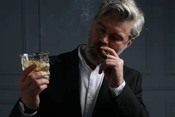 Handsome bearded man with glass of whiskey smoking cigar against dark grey background