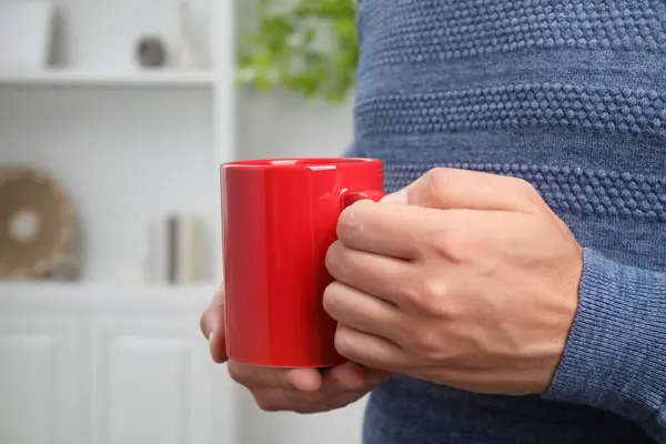 Man holding red mug at home, closeup. Mockup for design