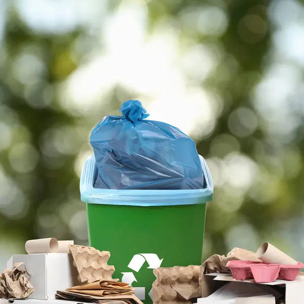 Waste bin with plastic bag full of trash surrounded by garbage on blurred background