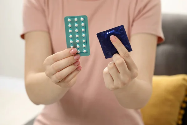 Woman holding condom and contraceptive pills on blurred background, closeup. Choosing birth control method