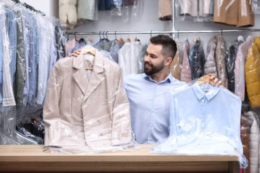 Kuru temizleme servisi. İçerideki tezgahta içinde plastik poşetler olan askıları tutan mutlu işçi.