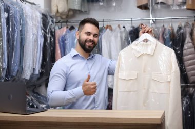 Kuru temizleme servisi. Naylon torba içinde askısı olan ve baş parmağını kapalı gişede gösteren mutlu işçi.