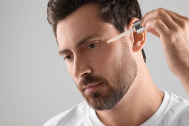 Yakışıklı bir adam yüzüne kozmetik serumu sürüyor açık gri arka planda, yakın plan.