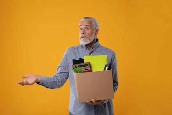 Confused unemployed senior man with box of personal office belongings on orange background