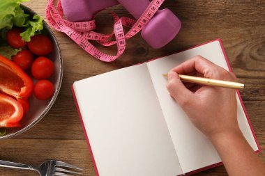 Ahşap masada, ürünlerle, mezuralarla ve dambıllarla beslenme planı geliştiren bir kadın.