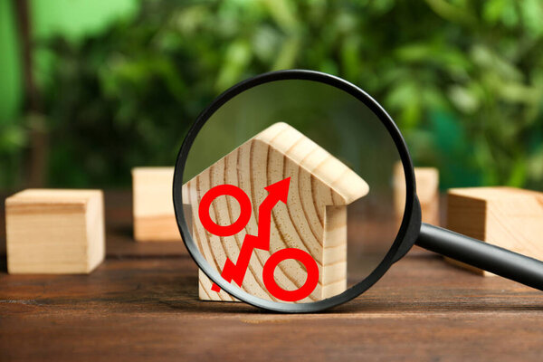 Рост ставки по ипотеке иллюстрируется знаком процента со стрелкой вверх. Модель дома на деревянном столе, вид через лупу