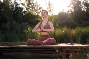 Güzel bir kadın, göl kenarındaki ahşap iskelede yoga minderinde Padmasana egzersizi yapıyor. Lotus pozu