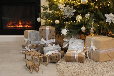 Evdeki şöminenin yanındaki güzel Noel ağacının altında birçok hediye kutusu ve dekoratif kızak var.