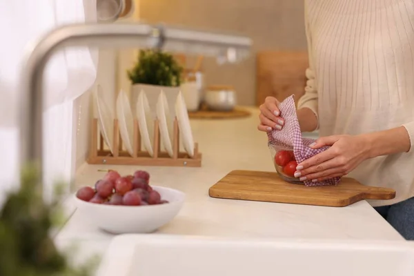 Woman packing bowl of fresh tomatoes into beeswax food wrap at white countertop in kitchen, closeup