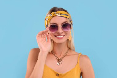 Açık mavi arka planda güneş gözlüklü gülümseyen hippi kadın portresi