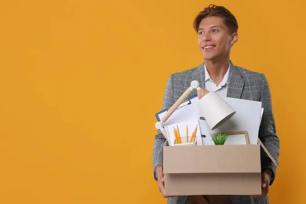 Happy unemployed young man with box of personal office belongings on orange background. Space for text