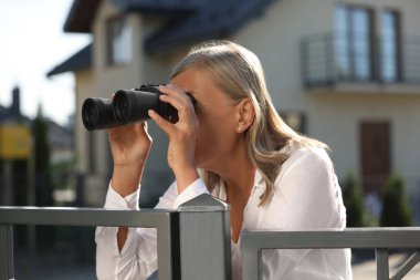 Concept of private life. Curious senior woman with binoculars spying on neighbours over fence outdoors clipart