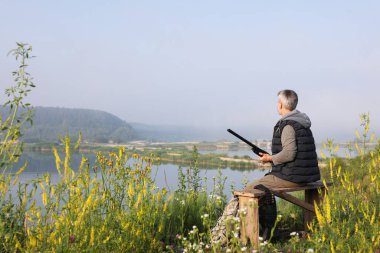 Avcı tüfekli bir adam gölün yakınındaki ahşap bankta oturuyor. Metin için boşluk