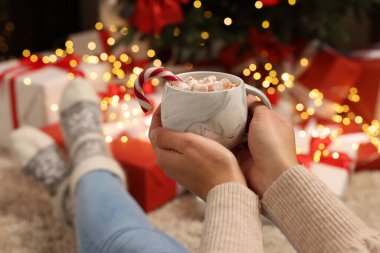 Bir kadın elinde bir fincan lezzetli kakao, marşmelov ve Noel hediyelerinin yanında şeker kamışı tutuyor. Yakın plan.