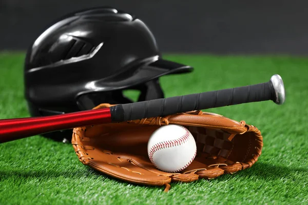 Baseball bat, batting helmet, leather glove and ball on green grass against dark background, closeup