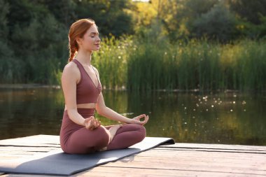 Güzel bir kadın yoga minderinde, göletin yanındaki tahta iskelede Padmasana antrenmanı yapıyor. Mesaj için yer var. Lotus pozu