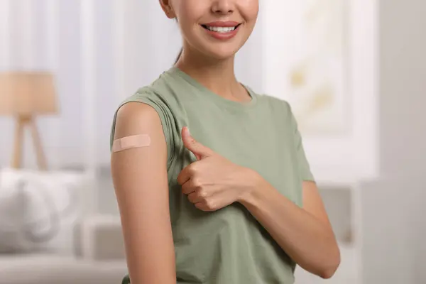 Woman with sticking plaster on arm after vaccination showing thumbs up at home, closeup