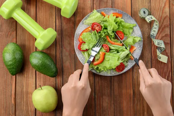 Healthy diet. Woman eating salad at wooden table with measuring tape and dumbbells, top view