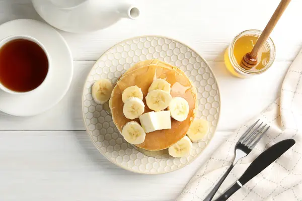 Delicious pancakes with bananas, honey and butter served with tea on white wooden table, flat lay