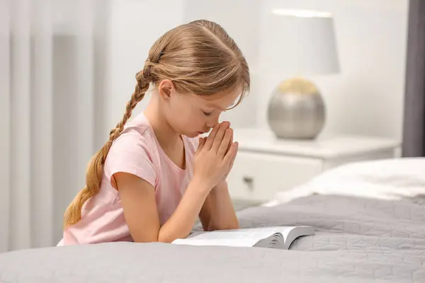 Girl holding hands clasped while praying over Bible in room