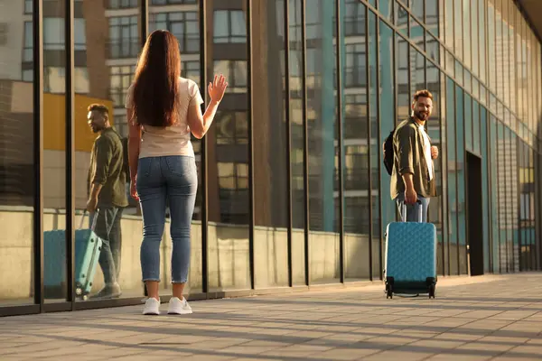 Long-distance relationship. Woman waving to her boyfriend with luggage near building outdoors