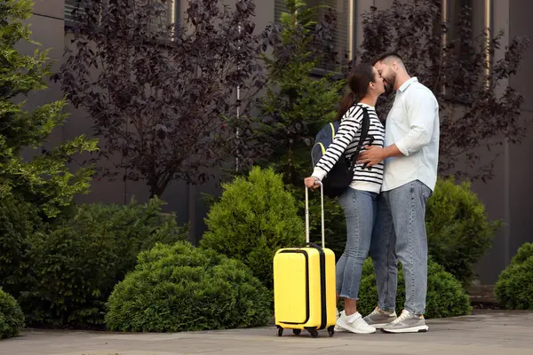 Long-distance relationship. Young woman with luggage kissing her boyfriend near building outdoors